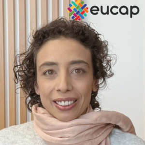 Grande plano de Raquel Tavares Lebre, uma mulher branca de cabelo curto encaracolado, com uma camisola branca e um cachecol cor-de-rosa. A Raquel sorri e tem um ar sereno.