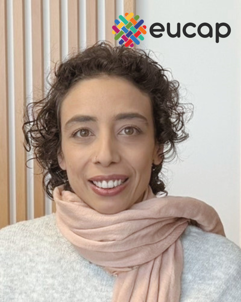 Grande plano de Raquel Tavares Lebre, uma mulher branca de cabelo curto encaracolado, com uma camisola branca e um cachecol cor-de-rosa. A Raquel sorri e tem um ar sereno.